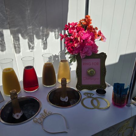 Sunlit outdoor bridal shower mimosa bar: glass carafes of colorful fruit juices, two chilled champagne bottles in ice wells, bright pink flower bouquet, framed “Ring for Champagne” sign, gold “Bride” headband and glitter ring cutouts.