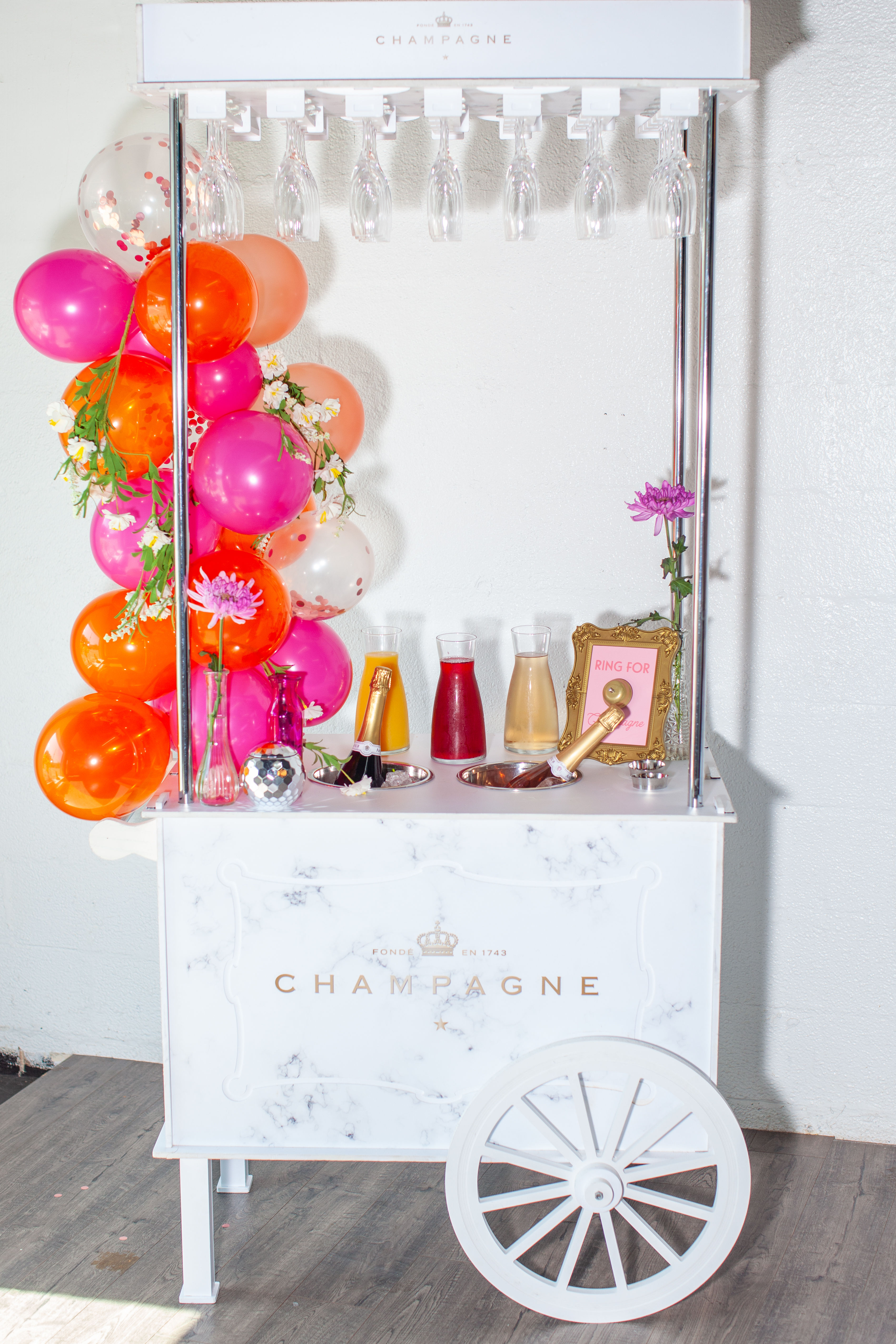 Festive white marble champagne cart party drink station with large wheel, hanging flutes, bright pink and orange balloon garland, champagne bottles, colorful carafes and floral accents against a white wall