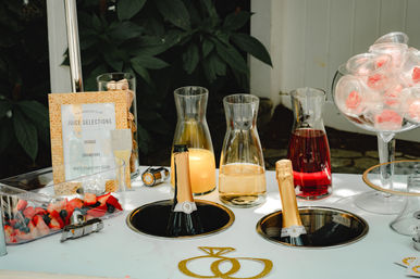 Brunch-ready outdoor mimosa bar with chilled champagne bottles in ice wells, carafes of orange, cranberry and peach juices, fresh berry platter and a glass bowl of pink macarons on a white table.