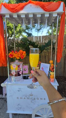 Hand holding a mimosa glass in front of a white champagne cart decorated with orange fabric, fresh oranges and vibrant flowers in a sunny backyard garden