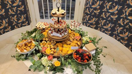 Indoor brunch display: tiered wooden stand and round table loaded with croissants, mini tarts, cupcakes, bowls of strawberries, blackberries and citrus halves, surrounded by greenery with decorative crane-pattern wallpaper in the background.