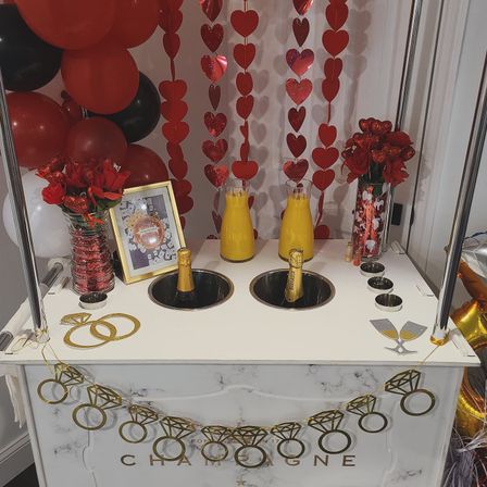 Party-ready indoor champagne cart decorated with red heart garlands and balloons, rose bouquets, two chilled champagne bottles in built-in wells and carafes of orange juice for mimosas.