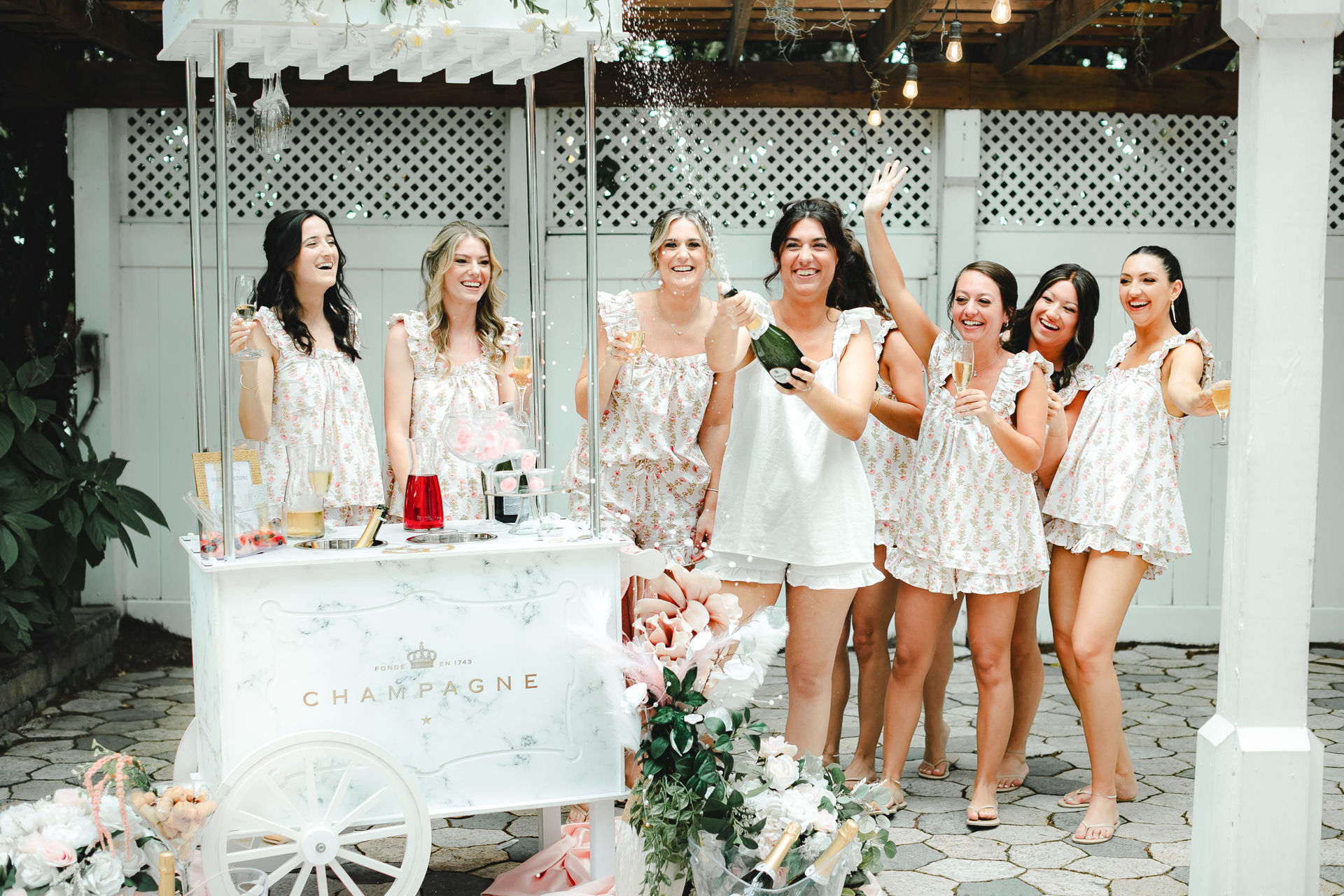 Bridal party in matching floral pajamas popping champagne and toasting by a white champagne cart on a sunny backyard patio — playful bachelorette/bridal shower celebration.