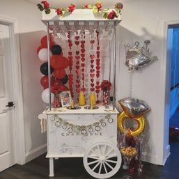 Chic white champagne cart in a home entryway decorated for a Valentine’s party with red heart garlands, red-black-white balloon column, hanging champagne flutes, juice carafes for mimosas, gold ring and diamond balloons, and floral accents.