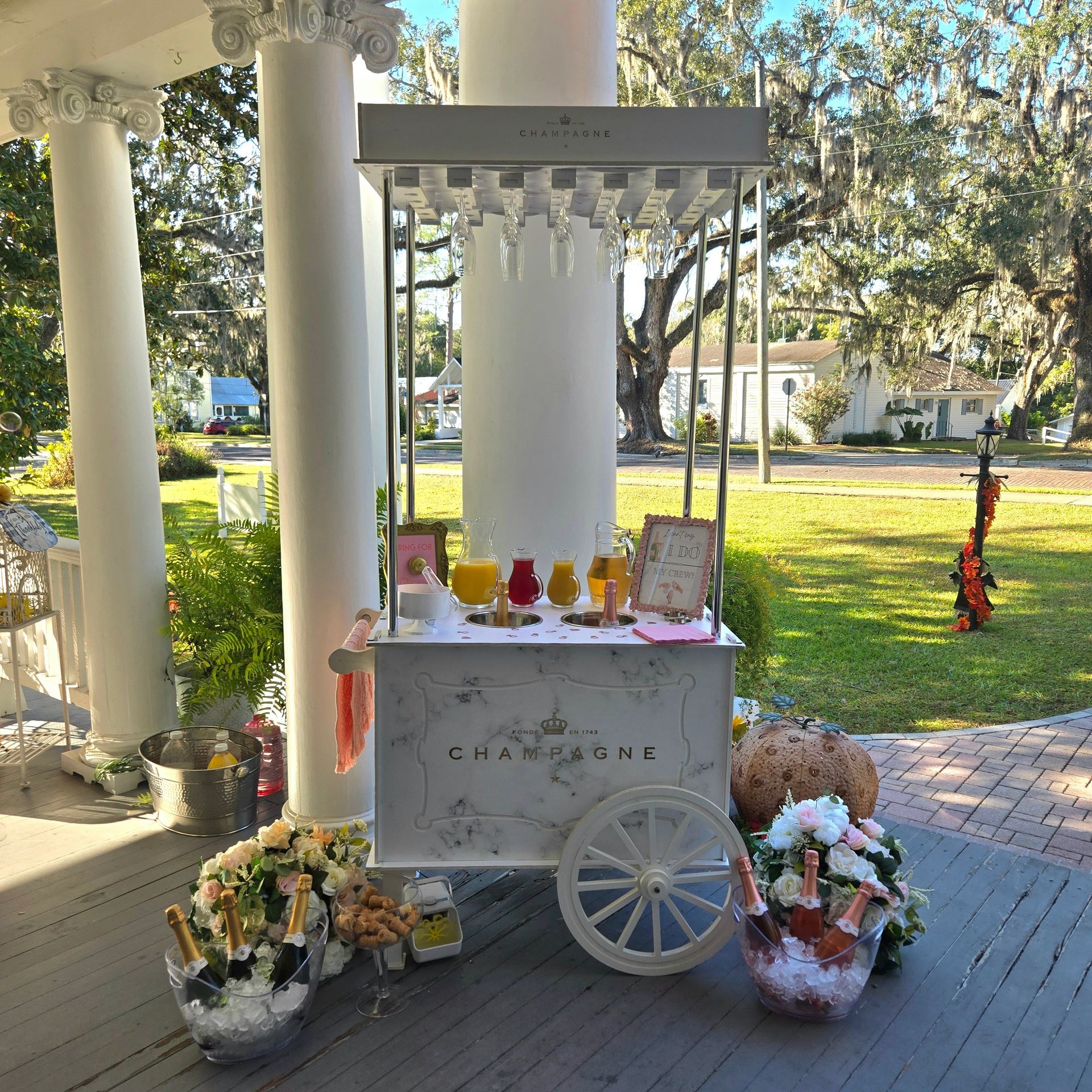 White vintage champagne cart on a columned southern front porch, stocked with hanging wine glasses, pitchers of orange and pink mimosas, buckets of champagne and rosé over ice, floral arrangements and pastries, with oak trees draped in Spanish moss on the lawn.