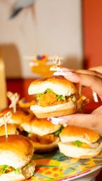 Hand holding a fried chicken slider with lettuce on a soft bun, colorful platter of mini sliders in the background