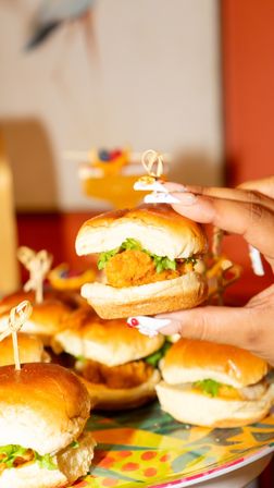 Hand holding a fried chicken slider with lettuce on a soft bun, colorful platter of mini sliders in the background