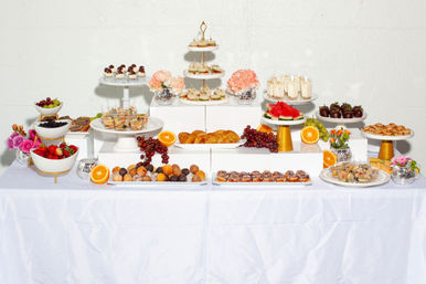 Elegant dessert buffet: tiered cake stand, cupcakes, mini parfaits, chocolate-covered strawberries, assorted donuts and pastries, fresh fruit (watermelon, grapes, strawberries, oranges) and floral accents on a white-and-gold display for a celebratory event