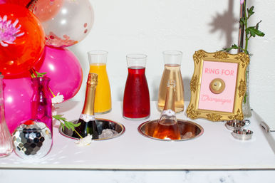 Brunch champagne and mimosa station for a party: two chilled champagne bottles in ice wells, three juice carafes (orange, red, rosé), pink balloons, decorative vases and a framed “Ring for Champagne” sign