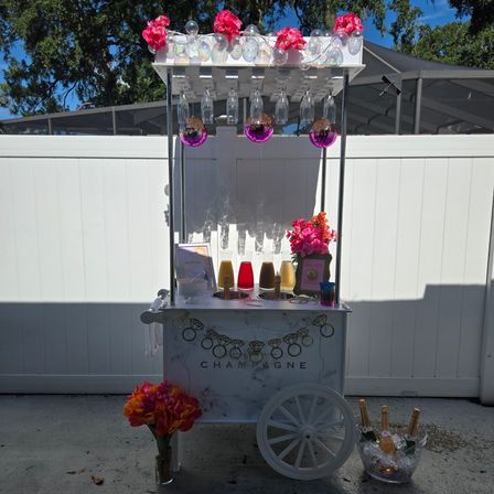 Backyard champagne cart decked with pink flowers, iridescent bubbles and mini disco balls, hanging flutes above colorful drink dispensers and an ice bucket of champagne bottles.