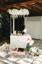 Chic outdoor white champagne cart on a sunlit garden patio, decorated with hanging wine glasses, cream-and-pink floral garlands, ice buckets of champagne bottles and soft pink draping for a wedding or event.
