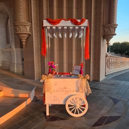 White champagne cart decorated with red fabric, gold bows and flowers on an ornate stone terrace at sunset — elegant wedding/event setup
