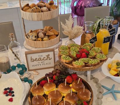 Beach-themed brunch spread with mini breakfast sliders on a wooden platter, two-tier basket of pastries, avocado toast rounds, fresh fruit and granola, juice carafes and pink foil balloon backdrop.