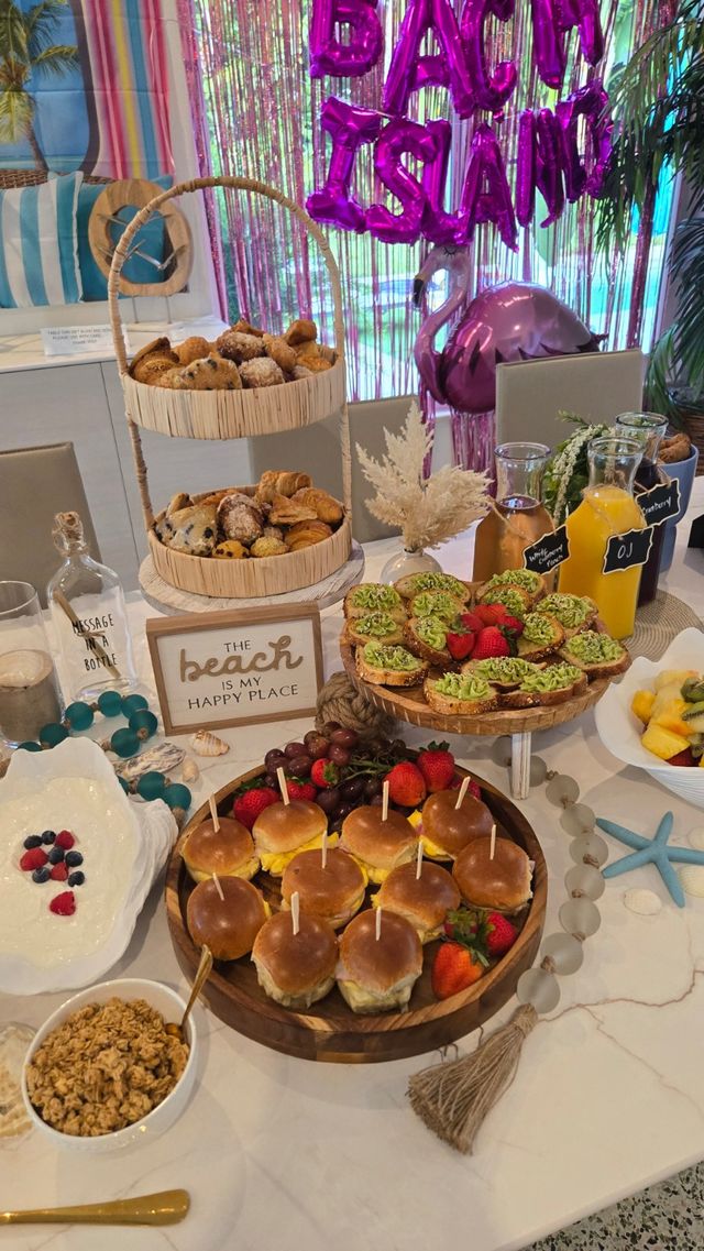 Beach-themed brunch spread with mini breakfast sliders on a wooden platter, two-tier basket of pastries, avocado toast rounds, fresh fruit and granola, juice carafes and pink foil balloon backdrop.