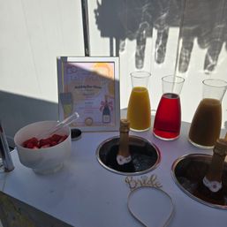 Bridal mimosa bar setup on a white table: three glass carafes of orange, red, and brown juices, two chilled champagne bottles in ice wells, a bowl of sliced strawberries, a framed bubbly-bar menu, and a gold "Bride" headband.