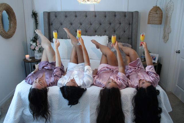Four friends in satin robes lying upside down on a bed with legs raised, toasting mimosas in a bright bedroom with a tufted headboard, white quilt, flowers and wicker decor — playful getting-ready moment.