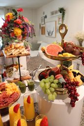 Vibrant home brunch spread on a kitchen island: two-tier gold fruit stand with grapefruit, grapes and macarons, charcuterie and pastries, green juice shots and strawberry-topped mimosas, bright floral centerpiece and cozy living room backdrop.
