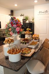 Bright home kitchen island brunch buffet with colorful floral centerpiece on a granite countertop, tiered trays of pastries, mini sandwiches, skewered appetizers and fresh finger foods, wicker barstools along the counter.