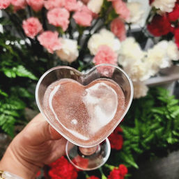 Close-up of a hand holding a heart-shaped glass of pink sparkling drink against a blurred bouquet of pink, white, and red flowers