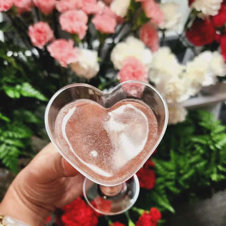 Close-up of a hand holding a heart-shaped glass of pink sparkling drink against a blurred bouquet of pink, white, and red flowers