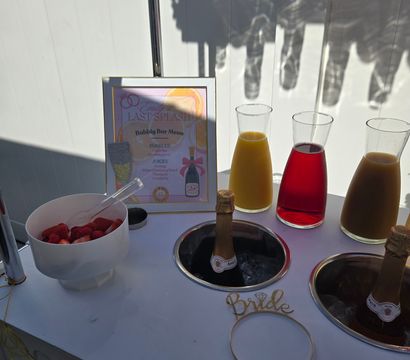 Bridal shower mimosa bar with chilled champagne bottles in ice wells, three glass carafes of orange, red and brown mixers, a bowl of strawberries and a framed bubbly bar menu on a white table.