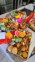 Colorful brunch grazing board on a marble kitchen island with chicken-and-waffles bites, biscuits, grapes, citrus halves, wraps, pastries and fresh flowers