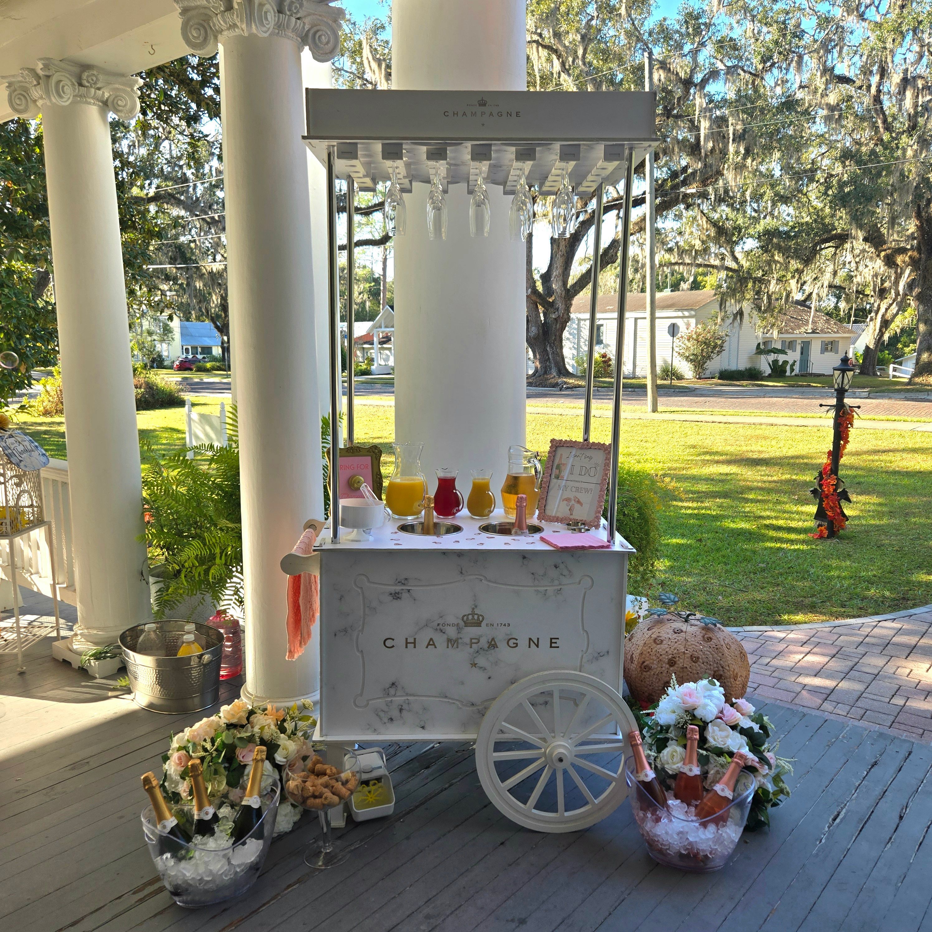 Festive white vintage champagne cart on a sunlit front porch with ice buckets of rosé and champagne, pitchers of mimosas, floral bouquets, and live oak trees draped with Spanish moss in the yard.