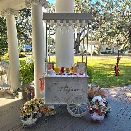 Festive white vintage champagne cart on a sunlit front porch with ice buckets of rosé and champagne, pitchers of mimosas, floral bouquets, and live oak trees draped with Spanish moss in the yard.