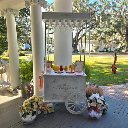 White champagne cart on a classic columned front porch, bottles on ice, glassware overhead, pitchers of mimosas and floral arrangements set for an outdoor wedding-style celebration beneath live oaks draped in Spanish moss.