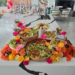 Bright party brunch spread on a marble kitchen island with sliders, canapés, grapes and citrus slices, vibrant pink and orange flowers, decorative balloons and metallic backdrop