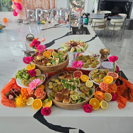 Bright party brunch spread on a marble kitchen island with sliders, canapés, grapes and citrus slices, vibrant pink and orange flowers, decorative balloons and metallic backdrop