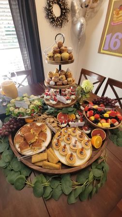 Party-ready indoor brunch spread on a wooden table with a three-tier stand of muffins and pastries, mini pancakes with syrup droppers, fruit cups and a mixed fruit bowl, slider sandwiches, cookies, orange juice pitcher, leafy garland centerpiece and silver balloons.