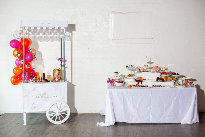 White champagne cart with hanging glasses and vibrant pink-orange balloons beside a white-draped dessert table piled with tiered pastries, fruit and floral accents against a white brick wall in a bright indoor party space.