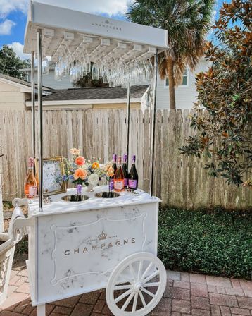 White mobile champagne cart with hanging flutes, rosé and sparkling bottles and a floral arrangement on a sunny backyard patio with a palm tree