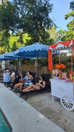 Poolside scene with black lounge chairs under scalloped blue umbrellas, people relaxing beside a white champagne cart draped in orange fabric and flowers, tropical trees and sunny backyard setting