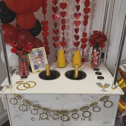 Romantic mimosa cart with two champagne bottles chilled in ice wells, carafes of orange juice, red heart garlands, red roses, black and red balloons and gold ring decorations.
