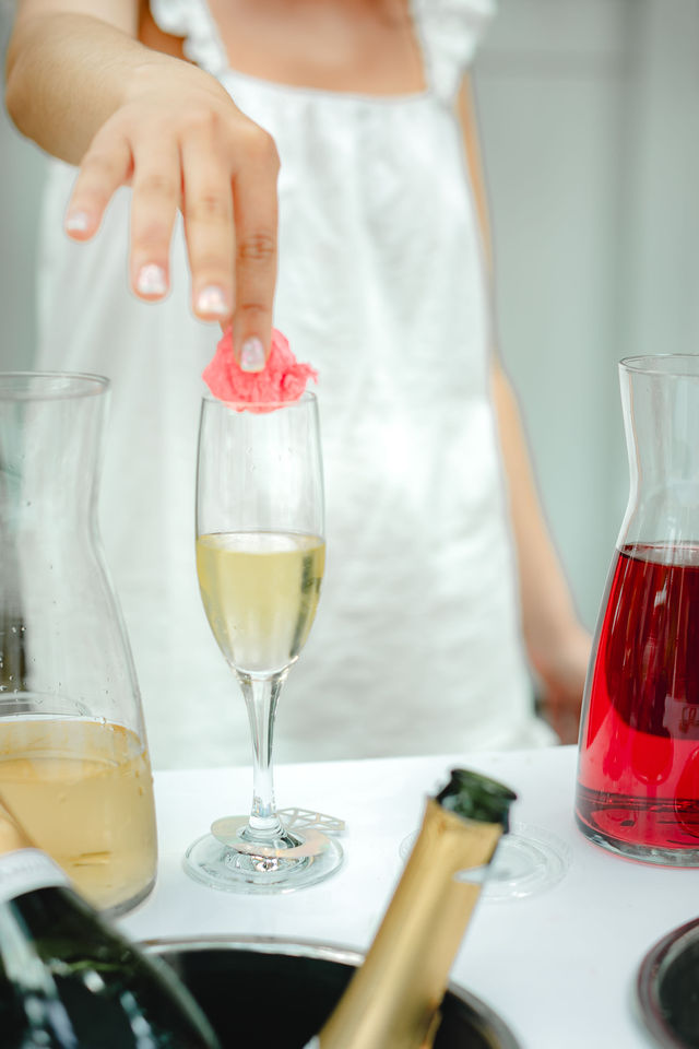 Hand placing a pink flower petal as a garnish on a sparkling champagne flute on a bright party table with carafes of red and pale drinks and a chilled bottle in an ice bucket