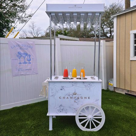 White vintage champagne cart with hanging flutes and three colorful carafes (red, yellow, orange) on artificial turf in a backyard beside a white fence and a "Last Toast" bridal shower banner.