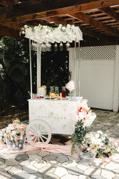 Sunlit outdoor champagne cart on a stone patio under a wooden pergola — white vintage cart with hanging wine glasses, floral garland, pink drapery and ice buckets with champagne and flowers, styled wedding drink station