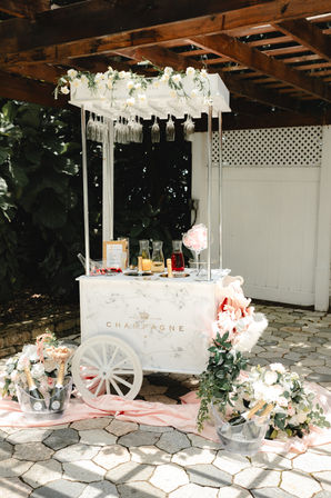 Sunlit outdoor champagne cart on a stone patio under a wooden pergola — white vintage cart with hanging wine glasses, floral garland, pink drapery and ice buckets with champagne and flowers, styled wedding drink station