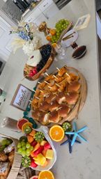 Brunch spread on a marble kitchen island with mini fried chicken sliders on a wooden board, yogurt and granola bowl with berries, colorful fruit platter (kiwi, pineapple, grapes, orange halves), croissants, gold utensils, decorative flowers and blue starfish accents.