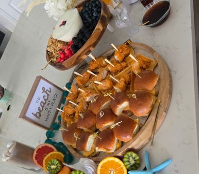 Brunch spread on a marble kitchen island with mini fried chicken sliders on a wooden board, yogurt and granola bowl with berries, colorful fruit platter (kiwi, pineapple, grapes, orange halves), croissants, gold utensils, decorative flowers and blue starfish accents.