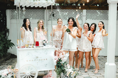 Outdoor bridal shower on a sunny patio: seven women in coordinating floral sleepwear laugh around a white champagne cart as one pops a bottle, holding glasses amid pink floral decor.