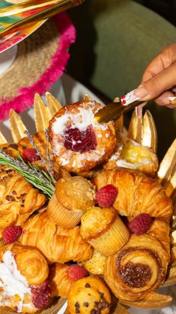 Close-up of a gold tray of assorted brunch pastries — jam-filled Danish with icing, buttery croissants, mini muffins and fresh raspberries, a hand using gold tongs to pick a pastry.