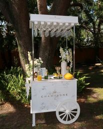 White marble-style champagne cart on a wooden wheel with hanging flutes, floral arrangements and carafes of champagne and orange juice set beneath large oak trees in a sunlit garden or backyard.