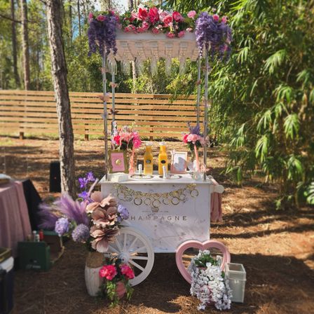 Outdoor mimosa/champagne cart in a pine grove — white vintage cart adorned with pink and purple floral garlands, hanging glassware, orange juice bottles, heart frame and bouquets for a garden party or wedding brunch.