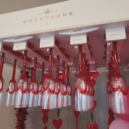 Row of upside-down champagne flutes with red stems hanging from a white rack labeled CHAMPAGNE, surrounded by red glitter hearts and pink balloons — festive Valentine’s Day bar decor
