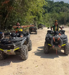 Two people on black ATVs stopped on a sunny dirt trail through lush tropical forest — one rider pointing ahead, the other posing for an off‑road adventure.