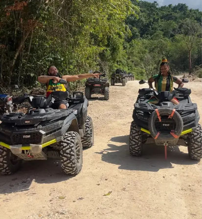 Two people on black ATVs stopped on a sunny dirt trail through lush tropical forest — one rider pointing ahead, the other posing for an off‑road adventure.