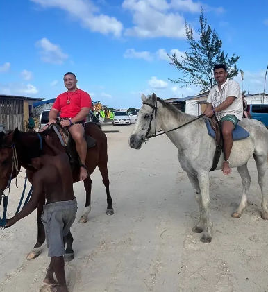 Two smiling riders on horseback trotting along a sandy coastal village street under a bright blue sky, with wooden buildings and parked cars in the background.